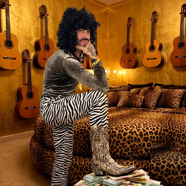 Er Richa, personaje de Ocurrensia, sentado con pantalón de cebra frente a una pared de guitarras